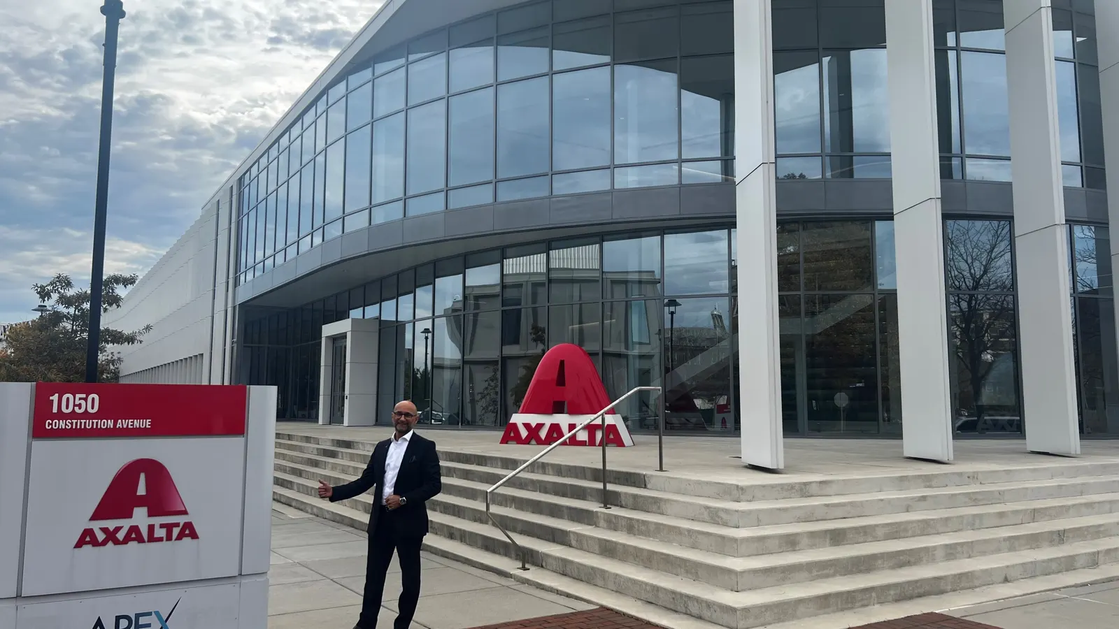 Enzo Santarsiero auf der Treppe vor einem Glasgebäude, daneben Schild und Skulptur mit dem Axalta-Logo